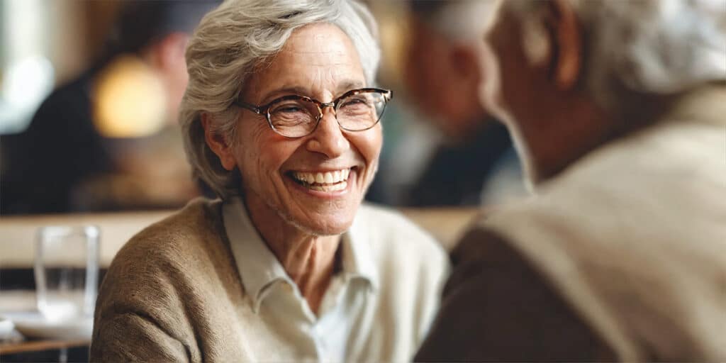 A person wearing dentures engaging in a lively and confident conversation with another individual, both smiling and enjoying the interaction in a relaxed, social setting.