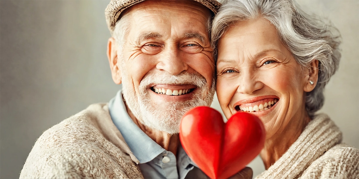 A senior couple smiling brightly, showcasing their healthy teeth, with a symbolic heart visible in the background, emphasizing the connection between oral health and heart wellness.