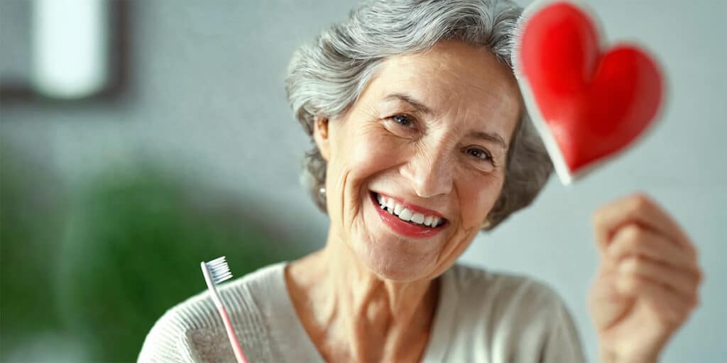 A senior woman smiling while holding a toothbrush, with a heart symbol prominently displayed in the foreground, representing the link between oral health and heart health.